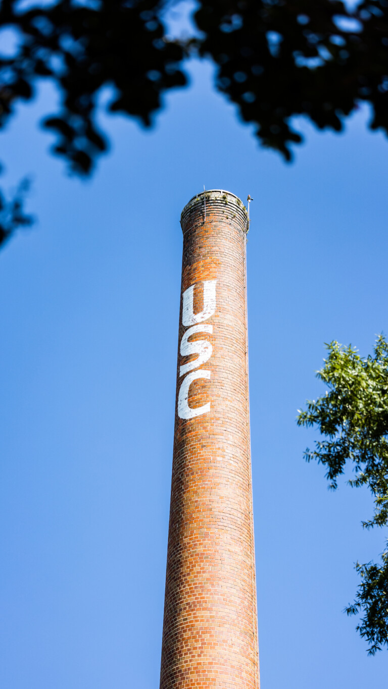 A tall brick chimney against a clear blue sky, featuring the letters 'USC' painted in white on its surface, with green foliage partially framing the image.