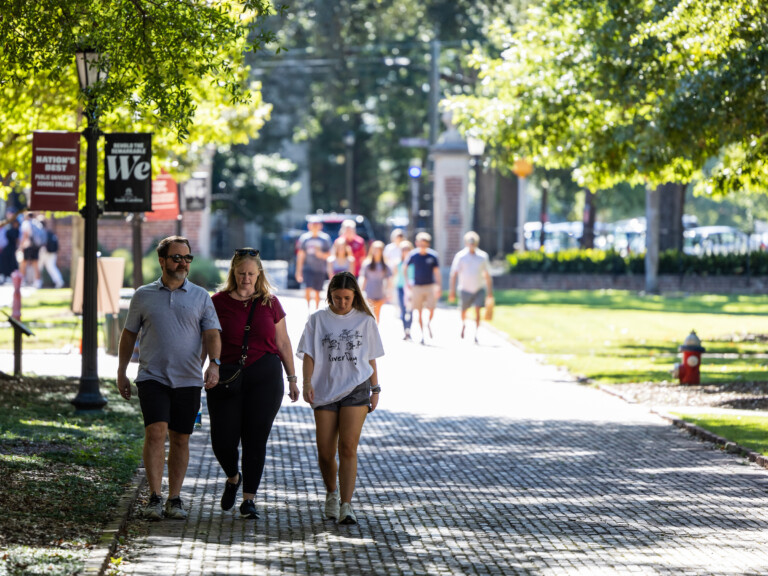 Three people, a man, a woman, and a young girl, walk hand-in-hand along a sunlit brick pathway lined with trees, while other pedestrians are visible in the background.