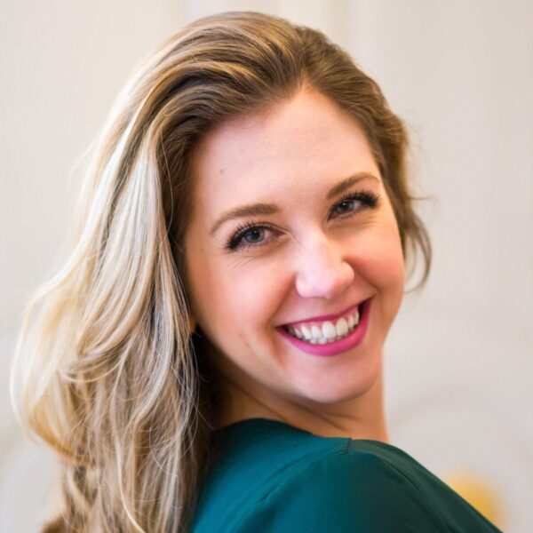 A smiling woman with long blonde hair, wearing a green top, poses with her head turned slightly to the side, against a softly blurred light background.