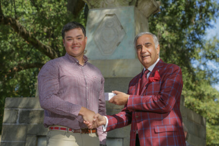 A young man in a checked shirt and beige pants shakes hands with an older man in a red plaid suit outside, with a stone monument visible behind them and greenery in the background.