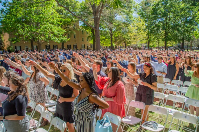 A large crowd of people outdoors, with many individuals raising their arms, likely in a celebratory or responsive gesture. The setting includes green trees and a building in the background, with rows of white chairs in the foreground. Attendees are dressed in various styles, suggesting a formal or semi-formal event.
