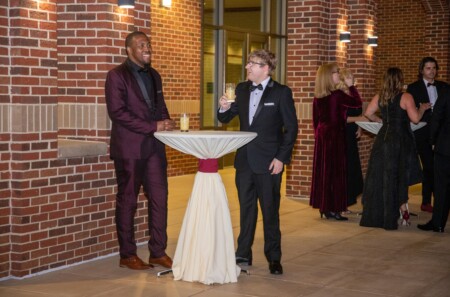 Two men in formal attire stand by a high table outdoors, one wearing a maroon suit and the other in a black tuxedo, both smiling and holding drinks; several other guests in evening wear can be seen in the background.