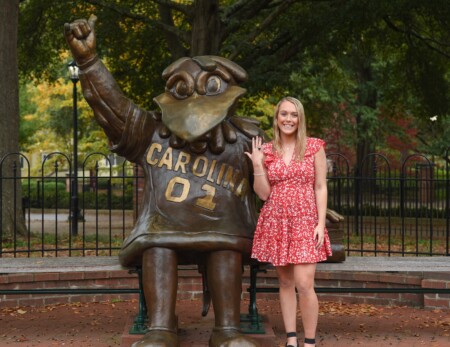 A woman in a red floral dress stands next to a large bronze statue of a university mascot, raising her hand with a smile. The background features trees with autumn foliage and a park-like setting.