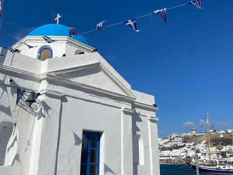 A close-up of a white building with a blue dome and a cross on top, decorated with Greek flags, against a clear blue sky, with a coastal town visible in the background.