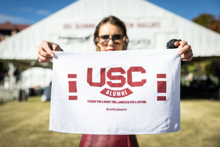 A person holding a white towel with red lettering that reads "USC ALUMNI" and the slogan "STUDENT FOR A SHORT TIME, GAMECOCK FOR A LIFETIME". A tent is visible in the background, indicating an outdoor event.