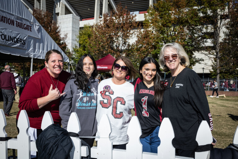 A group of four women and one girl pose in front of a fenced area during a tailgate event, with a large white tent in the background. One woman in a maroon sweater makes a hand gesture, while another wears a shirt with 'SC' on it, and the girl wears a jersey with 'CAROLINA' and the number '1'. The setting is lively with trees and a stadium structure visible in the background.
