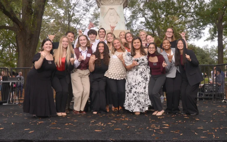 A diverse group of young adults poses for a photo on a stage in a park, with a decorative stone monument behind them. They smile and make hand gestures, wearing a mix of formal and casual attire, surrounded by trees with autumn foliage.