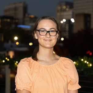 A young woman with glasses and a ponytail smiles at the camera, wearing an orange blouse, with city lights and a darkening sky in the background.