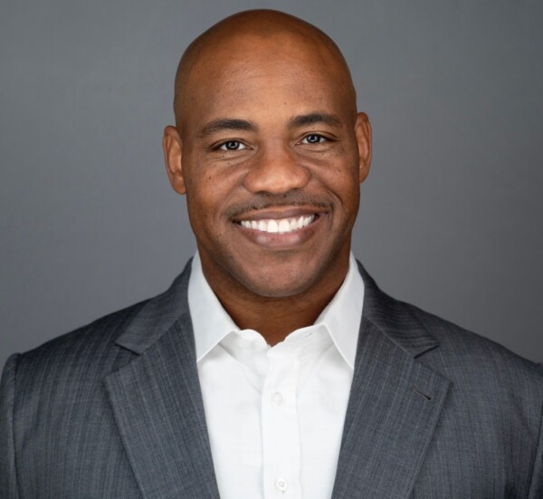 A smiling man with a bald head wearing a gray suit over a white shirt, posing against a neutral gray background.