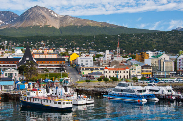 A picturesque harbor scene featuring several boats docked in the water, with colorful buildings lining the waterfront and a mountainous landscape in the background under a partly cloudy sky.