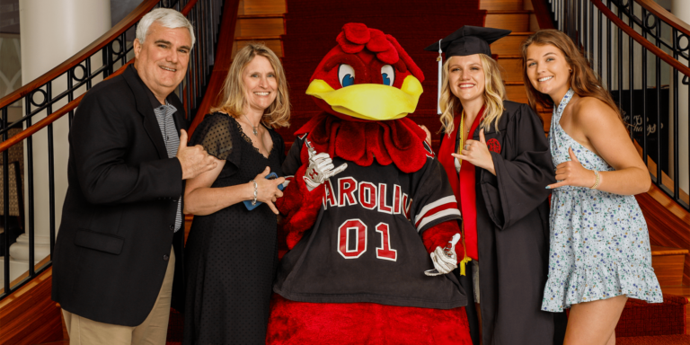 A family poses for a photo on a staircase with a mascot in a red and black outfit. The group consists of a man, a woman, a recent graduate wearing a cap and gown, and another woman. Everyone is smiling and making gestures with their hands, with the mascot's hand raised in a similar gesture.