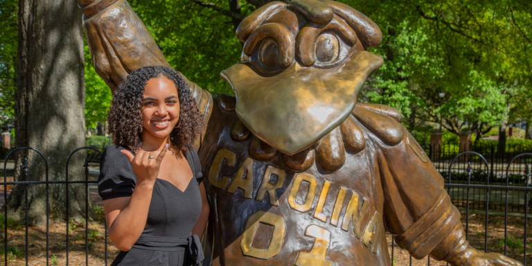 A young woman with curly hair smiles and gives a thumbs-up next to a large bronze statue of a mascot, wearing a shirt with 'CAROLINA' inscribed on it, set in a green park.