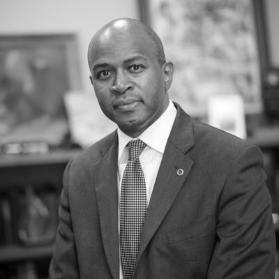 A professional man in a suit and tie sitting in an office setting, with books and framed pictures in the background.