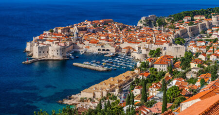 A panoramic view of Dubrovnik, Croatia, showcasing the old city with its iconic red-tiled roofs, medieval stone buildings, and the harbor filled with boats, set against a deep blue sea.
