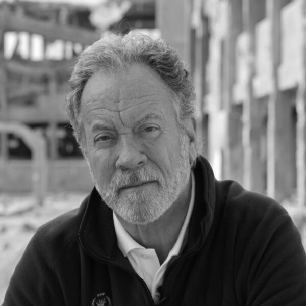 A mature man with gray hair and a beard, wearing a black jacket over a white shirt, looking directly at the camera with a serious expression. The background shows an industrial setting with blurred structures.