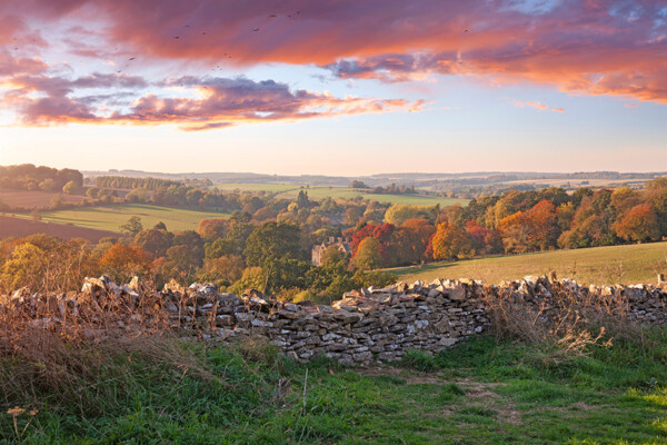 A scenic landscape featuring rolling hills and a stone wall in the foreground, with vibrant autumn foliage in shades of orange and red, under a colorful sky at sunset.