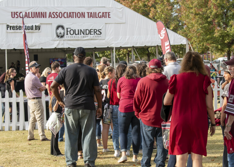 A line of people waiting at the USC Alumni Association tailgate event, with many wearing game day attire in the school's colors, in front of a large tent that displays the event's name and sponsors.