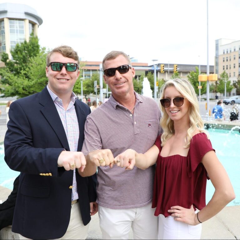 Three people standing in front of a fountain, smiling and showing their hands with rings. The man on the left wears sunglasses and a blazer over a checked shirt, the man in the middle wears a striped shirt and sunglasses, and the woman on the right wears a maroon top and sunglasses.