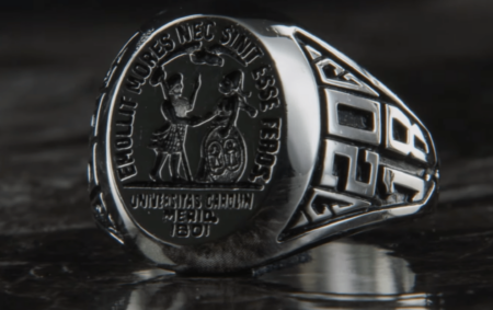 A silver university ring featuring intricate engravings, including a depiction of two figures, with Latin inscriptions and 'Universitas' above them, on the front of the ring, and decorative designs along the sides.