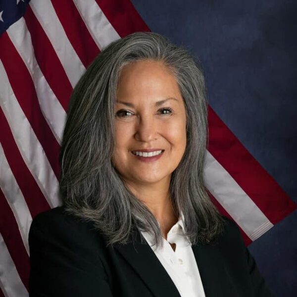 A woman with long, gray hair smiles confidently in front of a backdrop featuring the American flag. She is wearing a black blazer and a white blouse, projecting a professional appearance.