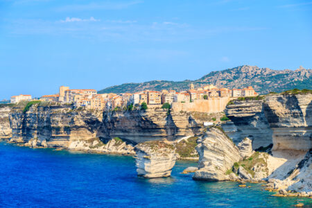 A coastal view of the town of Bonifacio in Corsica, featuring multi-colored buildings on cliff edges, white limestone cliffs, and a deep blue sea under a clear sky.