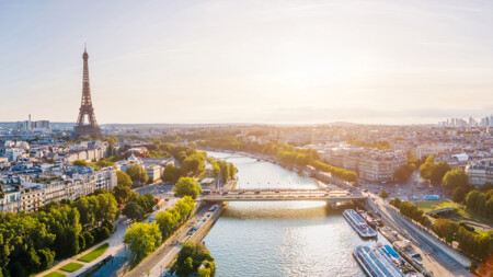 Aerial view of the Eiffel Tower in Paris, surrounded by lush green trees, the Seine River flowing beneath a bridge, and a vibrant cityscape under a golden sunset.