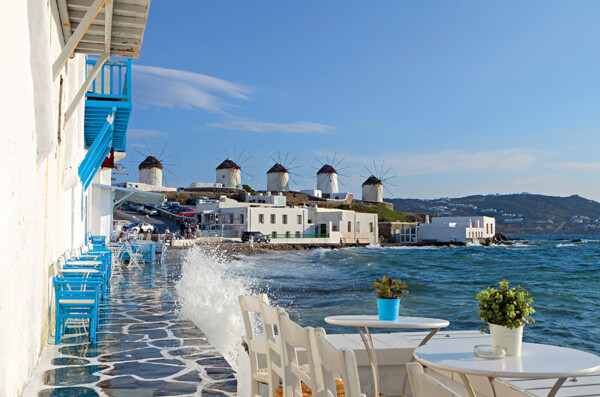 A seaside view in Mykonos featuring whitewashed buildings with blue accents, blue chairs along a stone path, and multiple traditional windmills on a hill in the background, with waves splashing on the shore and a small table with a potted plant in the foreground.