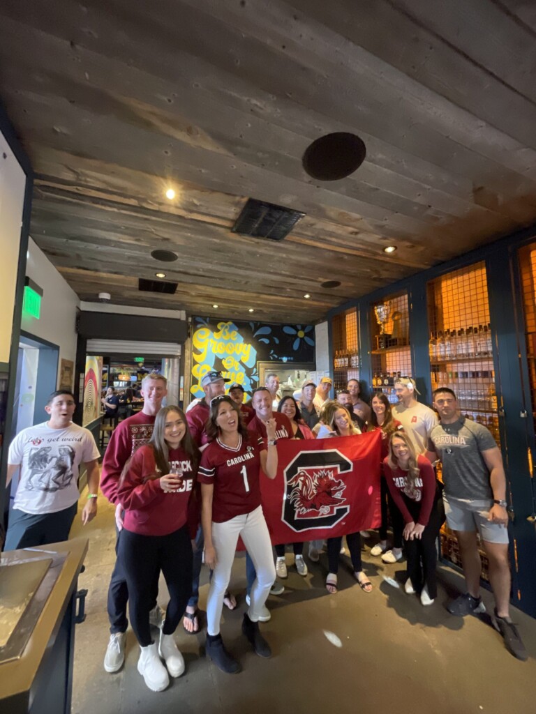 A group of people gathered in a lively indoor setting, wearing garnet and black clothing with the South Carolina Gamecocks logo. They are holding a Gamecocks flag and posing happily for the photo, with a colorful mural and wooden ceiling in the background.