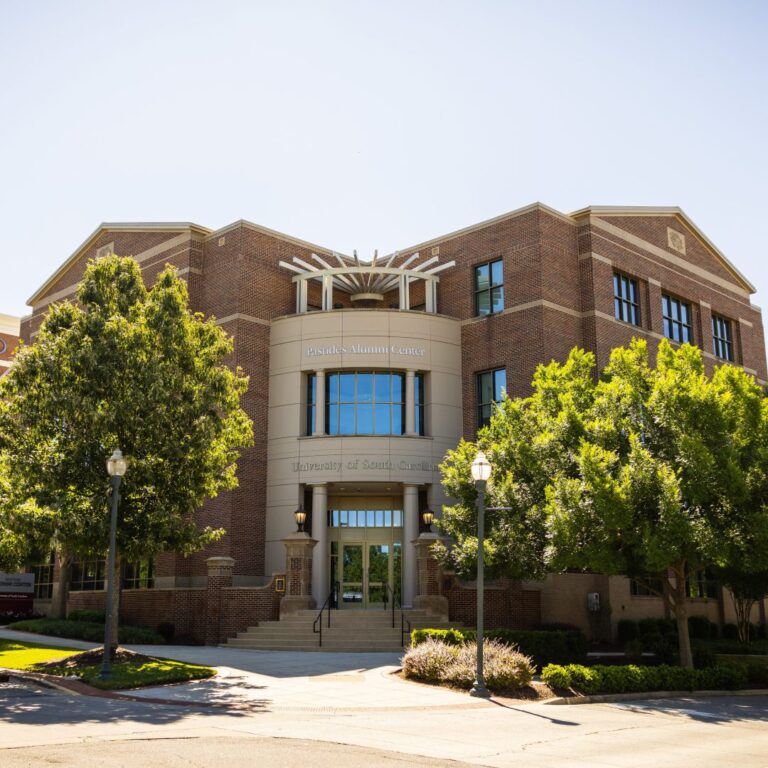 A view of the Psides Alumni Center at the University of South Carolina, featuring a brick facade, large glass windows, and landscaping with trees and shrubs, under a clear blue sky.