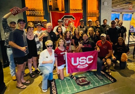 A large group of people gathered in a venue, many wearing sports jerseys and holding a flag that reads 'USC ALUMNI'. The atmosphere is festive, with decorations likely related to a sports event, and a prominent Gamecock logo on the floor mat.