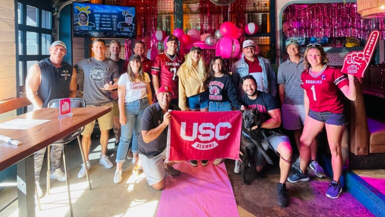 A group of USC alumni poses for a picture in a decorated venue, holding a USC flag. The background features pink balloons and streamers, and a scoreboard displaying game stats is visible on a screen. The group includes both men and women, some wearing USC gear, with one person holding a foam finger, and a dog sitting at their feet.
