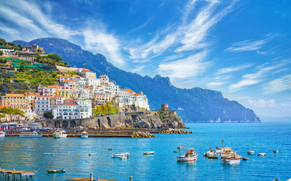A picturesque coastal scene featuring colorful buildings on a hillside overlooking a blue sea, with small boats floating near a rocky shoreline and mountains in the background under a partly cloudy sky.