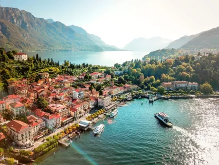 Aerial view of a picturesque lakeside town with colorful buildings, lush greenery, and mountainous backdrop, showcasing boats moored along the water's edge under a clear sky.