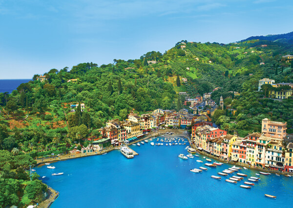 A scenic view of Portofino, Italy, showcasing colorful waterfront buildings along the harbor, surrounded by lush green hills and a bright blue sky, with several boats anchored in the calm water.