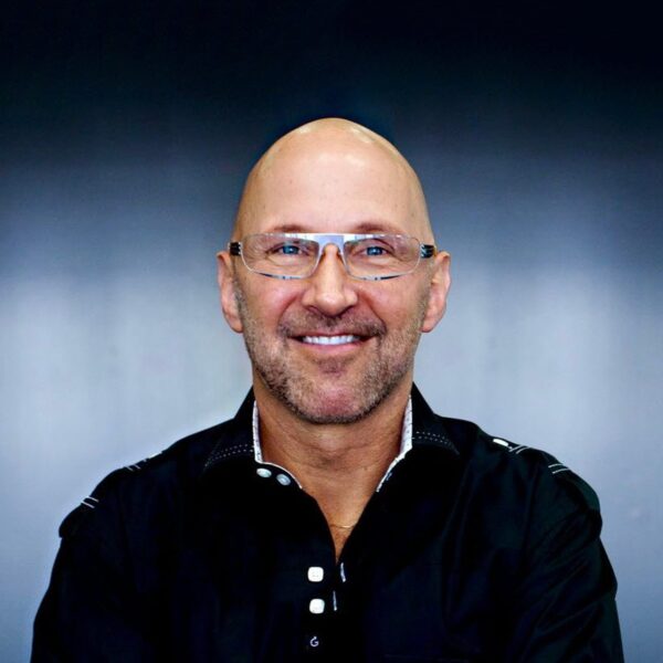 A smiling man with a bald head and glasses, wearing a black shirt with a decorative collar, posed against a blurred gray background.