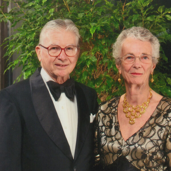 An elderly couple posing together, the man in a tuxedo with a bow tie and glasses, and the woman wearing an elegant black and gold outfit with a statement necklace, against a background of lush green foliage.
