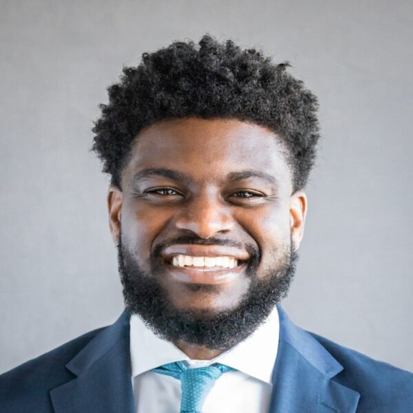 A smiling man with short, curly black hair and a full beard, wearing a dark suit and light blue tie, against a gray background.