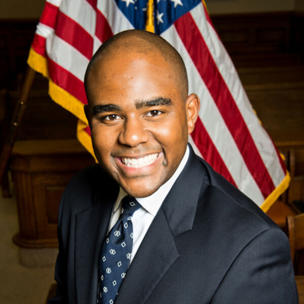 A smiling man in a dark suit and patterned tie poses for a photo in front of American flags, with a wooden interior visible in the background.