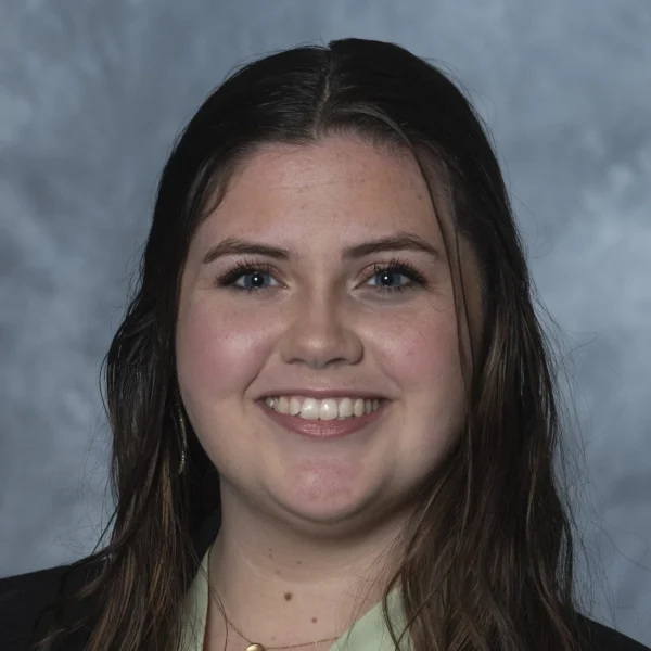 A young woman with long, dark hair smiles at the camera, wearing a black blazer over a light green top, with a soft gray background.
