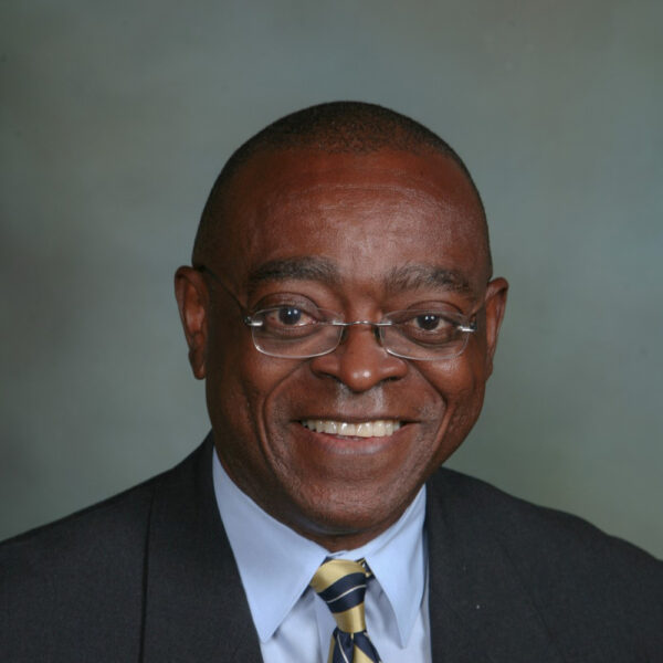 A smiling man wearing glasses, a dark suit, and a light blue shirt with a striped tie, set against a muted gray background.