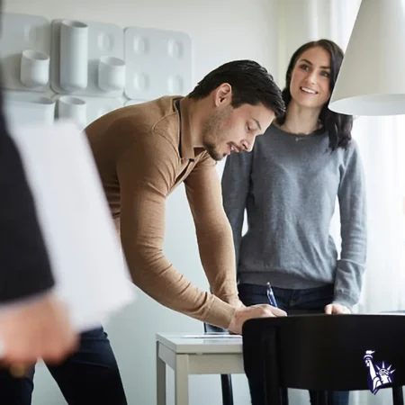 A man in a brown sweater is leaning over a table, writing on a piece of paper with a pen, while a woman in a gray sweater stands nearby, smiling at him. The background features modern wall decor and natural lighting from a window.
