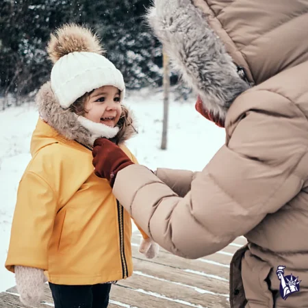 A smiling child in a yellow winter jacket and a white knitted hat is being helped by an adult, who is wearing a brown puffer coat, to adjust the jacket in a snowy outdoor setting.