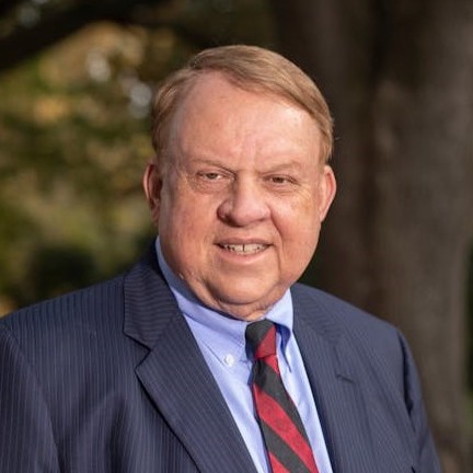 A middle-aged man with light brown hair, wearing a dark pinstripe suit, blue dress shirt, and a striped tie in red and black, poses outdoors against a blurred natural background that suggests a park or garden setting.