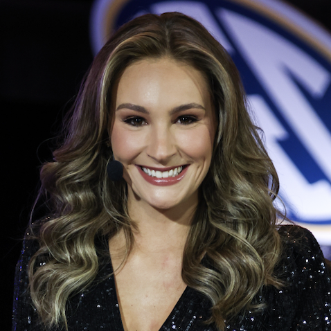 A woman with long, wavy hair and a bright smile, wearing a sparkly black outfit, stands in front of a logo backdrop, appearing as a host or presenter.