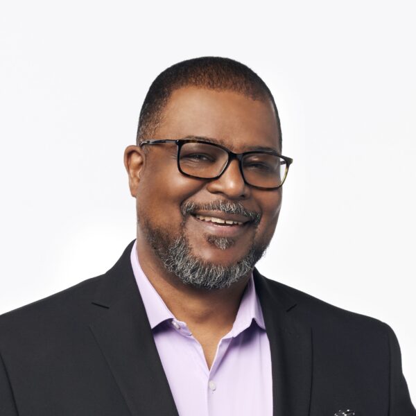 A smiling man with glasses, short black hair, and a gray beard, wearing a black blazer over a light purple shirt, against a plain white background.