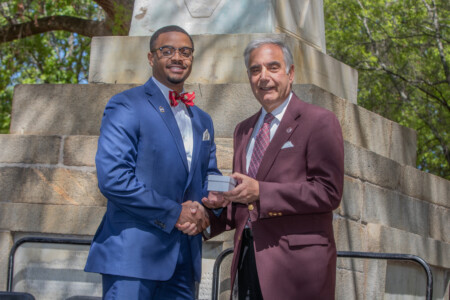 A young man in a blue suit and red bow tie shakes hands with an older man in a maroon blazer, both smiling. The older man is holding a small box, and they are standing in front of a stone monument with trees in the background.