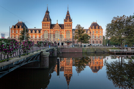 A historic building with ornate architecture reflecting in a calm body of water, surrounded by lush trees and colorful flower arrangements in the foreground, under a clear blue sky.