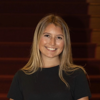A smiling young woman with long, straight blonde hair, wearing a black t-shirt, is standing in front of wooden steps.