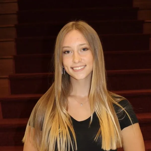 A smiling young woman with long, straight blonde hair stands in front of a staircase with red carpeting, wearing a black top and a small necklace.