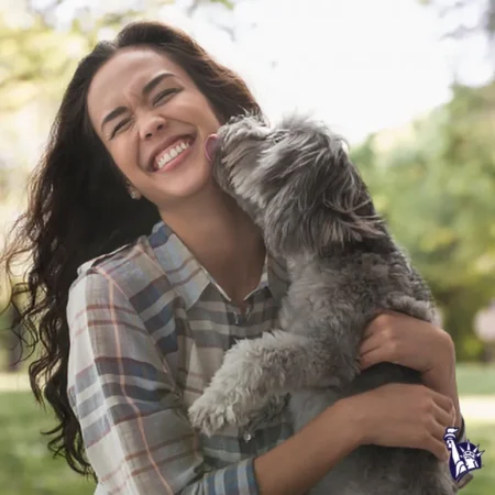 A young woman with long, curly hair smiles broadly while holding a small, fluffy dog that is licking her face, set in a green outdoor environment.
